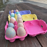 a pink 6-egg pulp unprinted carton with colored, speckled eggs in the cell. There the same carton in yellow and purple in the distance.